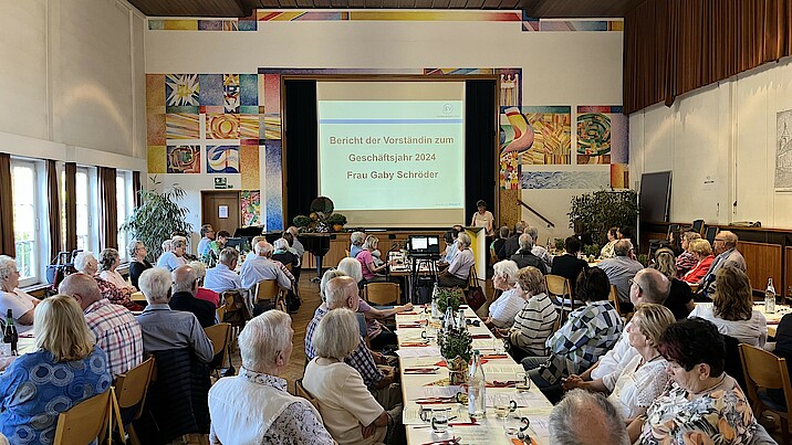 Viele Menschen sitzen an Tischreihen in einem Saal und blicken auf eine Präsentation auf der Bühne