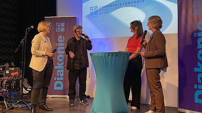 Vier Personen stehen vor einer Leinwand um einen Stehtisch herum. Es handelt sich um eine Interviewsituation.