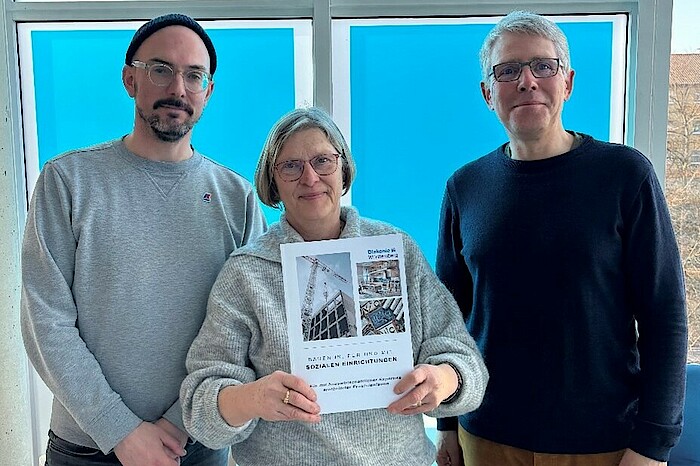 Zwie Männer und mittig eine Frau sind vor einem blauen Fenster zu sehen und die Frau hält eine Publikation in den Händen.