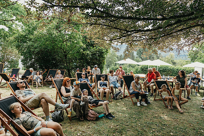 Menschen sitzen auf Liegestühlen im Freien unter Bäumen.