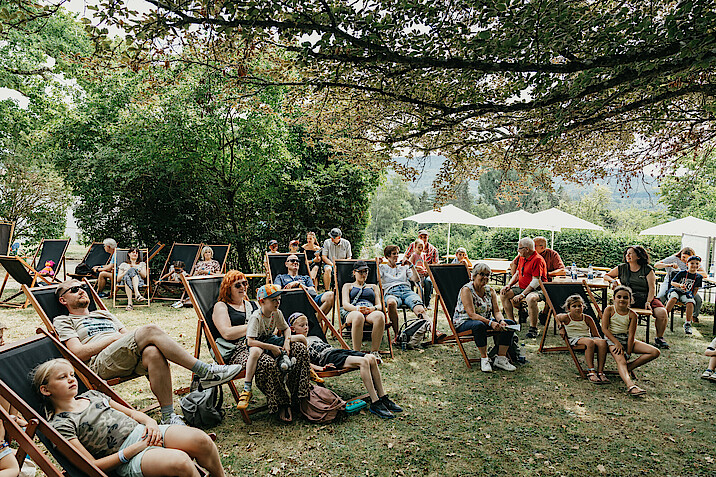 Menschen sitzen auf Liegestühlen im Freien unter Bäumen.