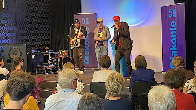 Drei Männer stehen auf einer Bühne und singen in Mikrofone. Davor sitzt das Publikum und schaut auf die Bühne.
