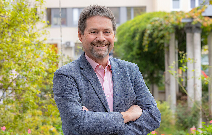 Ralf Horndasch steht lächelnd und mit verschränkten Armen da, er trägt ein graues Sakko.