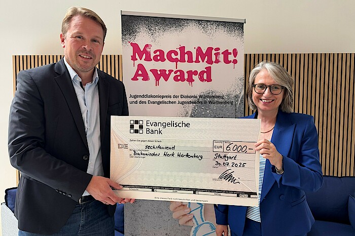 Oberkirchenrätin Dr. Annette Noller und Sebastian Schneider, Regionaldirektor Stuttgart bei der Evangelischen Bank, stehen vor einem Aufsteller des Jugenddiakoniepreises, halten einen Scheck in der Hand und lachen in die Kamera.