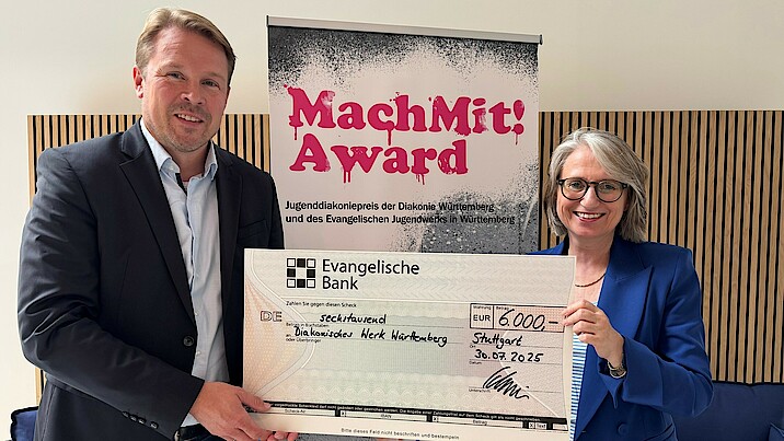 Oberkirchenrätin Dr. Annette Noller und Sebastian Schneider, Regionaldirektor Stuttgart bei der Evangelischen Bank, stehen vor einem Aufsteller des Jugenddiakoniepreises, halten einen Scheck in der Hand und lachen in die Kamera.