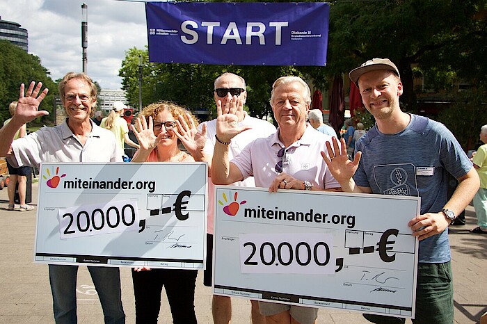 Fünf Menschen halten zwei Spendenschecks über 20000 Euro in die Kamera