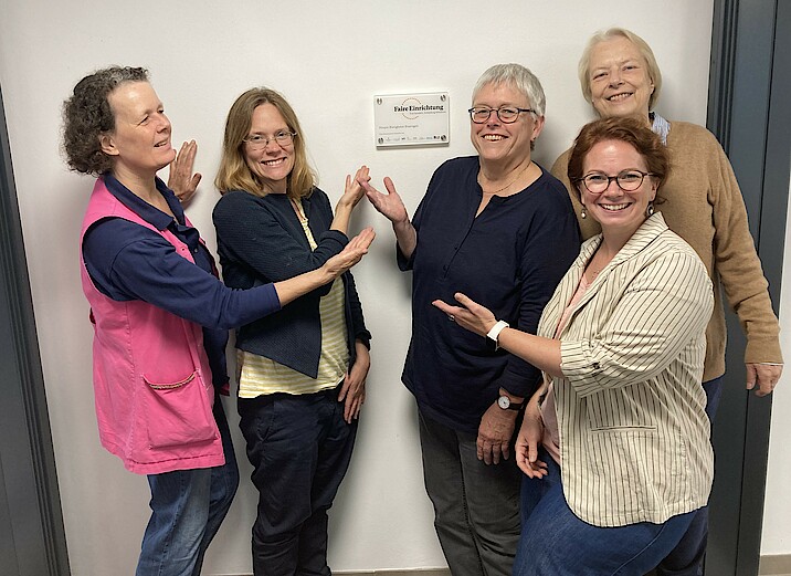 Fünf Frauen stehen lächelnd vor einer Wand und zeigen auf ein Türschild mit der Aufschrift "Faire Einrichtung"