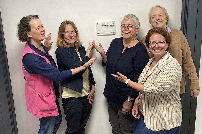 Fünf Frauen stehen lächelnd vor einer Wand und zeigen auf ein Türschild mit der Aufschrift "Faire Einrichtung"