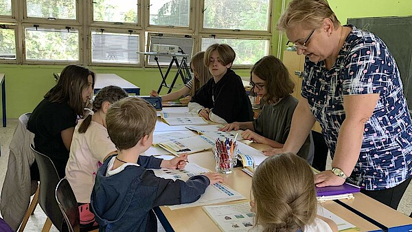 Sechs Kinder und eine Lehrerin stehen und sitzen an einem Tisch und lernen.