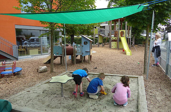 Drei Kinder spielen im Sandkasten, über den ein grünes Segel gespannt ist.
