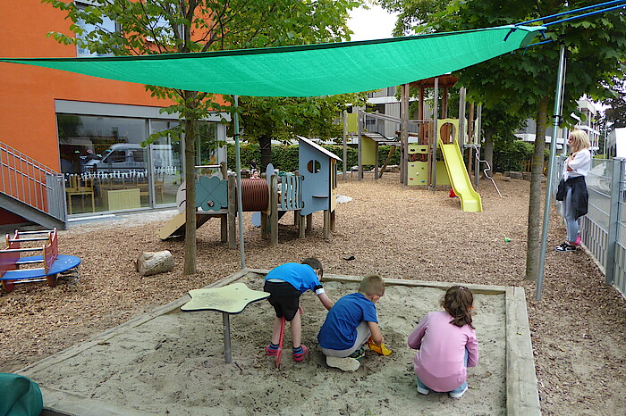 Drei Kinder spielen im Sandkasten, über den ein grünes Segel gespannt ist.