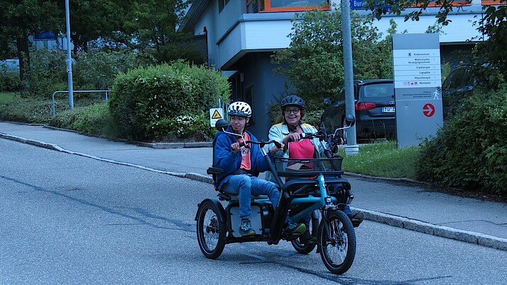 Zwei Personen fahren lächelnd in einem Tandem die Straße hinunter.