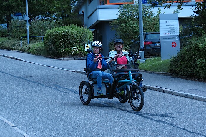 Zwei Personen fahren lächelnd in einem Tandem die Straße hinunter.
