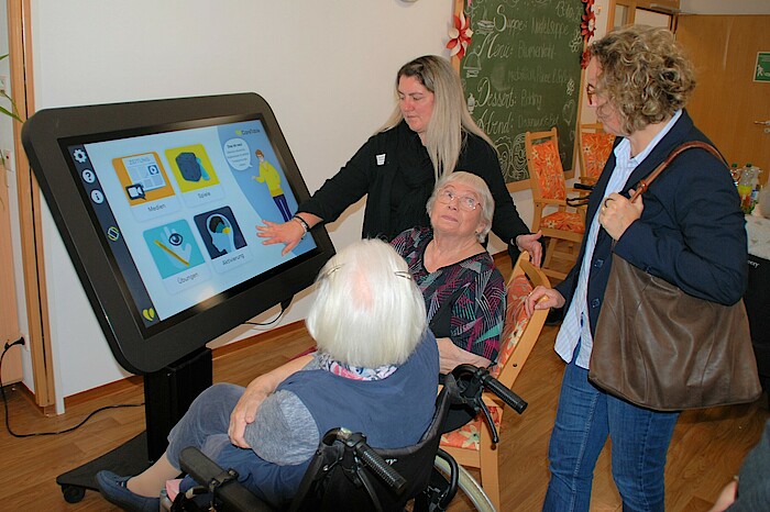 Vier Frauen sitzen und stehen von einem großen Bildschirm, eine Frau drückt auf ein Symbol.