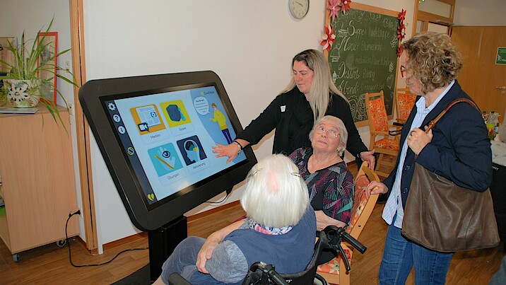 Vier Frauen sitzen und stehen von einem großen Bildschirm, eine Frau drückt auf ein Symbol.