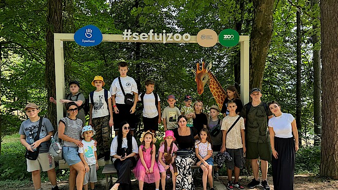 Eine Gruppe von Kindern mit Erwachsenen steht am Eingang eines Zoos im Grünen.