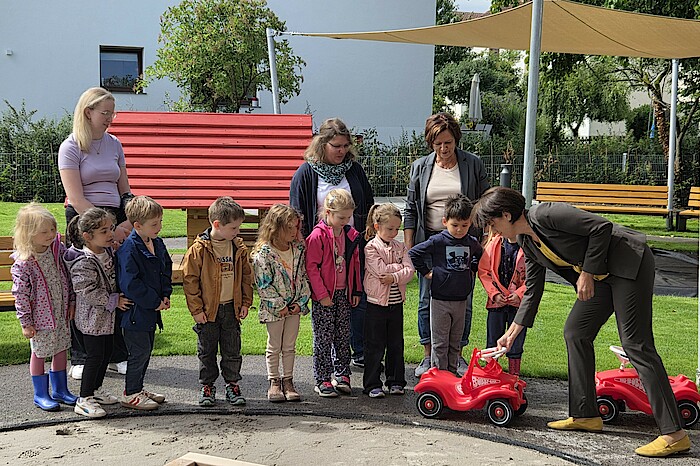Mehrere Kinder und Erwachsene stehen auf einem Spielplatz. Eine Frau zeigt den Kindern ein rotes Auto und alle schauen aufmerksam darauf