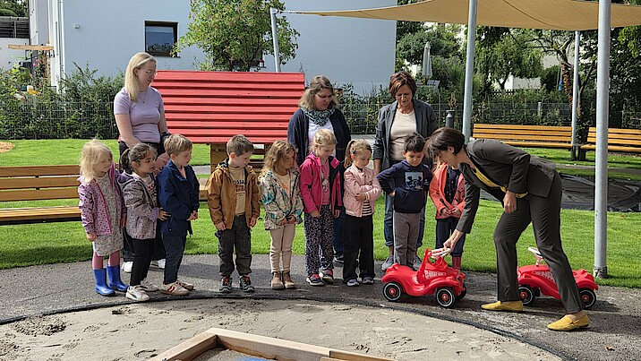 Mehrere Kinder und Erwachsene stehen auf einem Spielplatz. Eine Frau zeigt den Kindern ein rotes Auto und alle schauen aufmerksam darauf