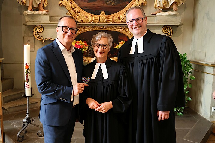Drei lächelnde Personen stehen in einer Kirche und schauen in die Kamera